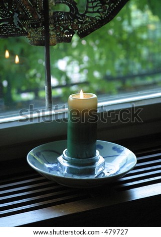 Atmospheric low-light shot of lit candle in the window at dusk with reflections of the flame in the window glass.