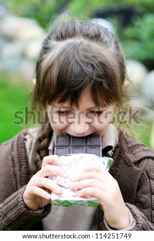 Little cute girl biting big tablet of chocolate, focus on girl's face