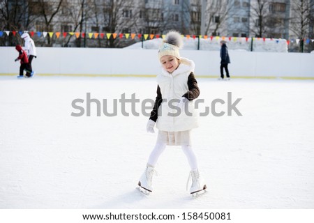 Adorable little girl in the white clothes and hat on the ice rink