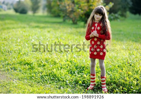 Adorable kid girl in red sweater dress and stripe rainboots having fun with bird feathers outdoors in the sunny autumn day