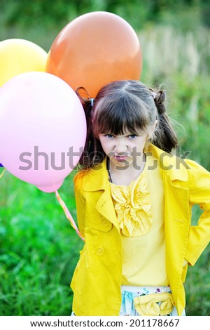 Beauty brunette kid girl with funny angry face in yellow jacket with bunch of colorful balloons