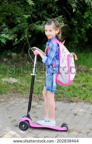 Beauty school age  kid girl in plaid shirt and jeans skirt with rucksack rides on her pink scooter from the school and listen to the music with her cell phone