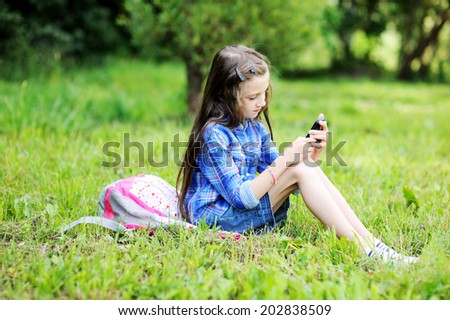 Beauty  fashion brunette school age kid girl in the blue plaid shirt, jeans skirt and yellow scarf writing in her notepad outdoor on the playground