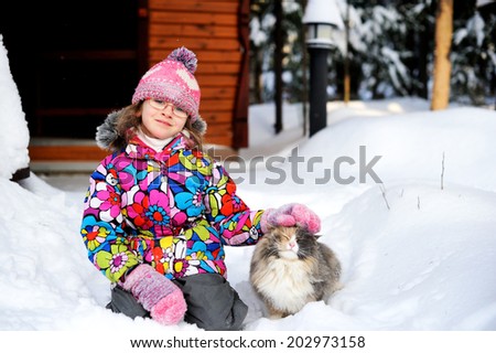 Beauty little girl in colorful winter jacket and hat plays with a fury cat in deep snow on nice winter day
