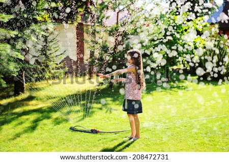 Nice kid girl with long hair has fun with garden sprinkler playing with water splashes in the backyard on a sunny hot summer  evening.