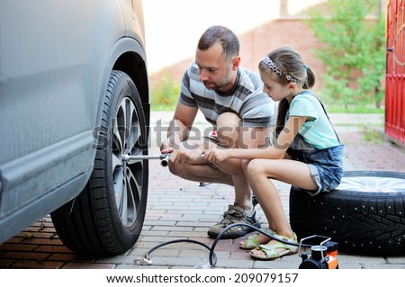 Cute little girl helps her father to change wheel on their family car on warm day in the yard