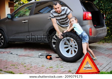 Cute little girl helps her father to change wheel on their family car on warm day in the yard