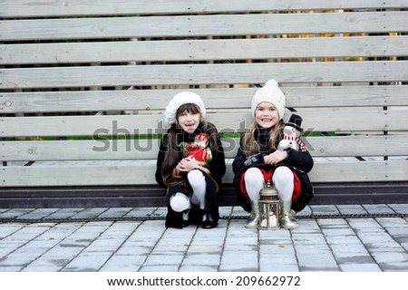 Two adorable kid girl friends in white hats and pattern black jackets has fun outdoor  at the winter day