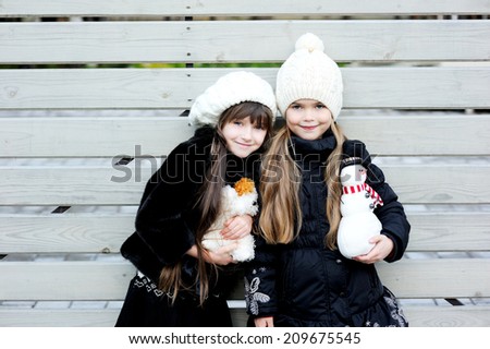 Two adorable kid girl friends in white hats and pattern black jackets has fun outdoor at the winter day