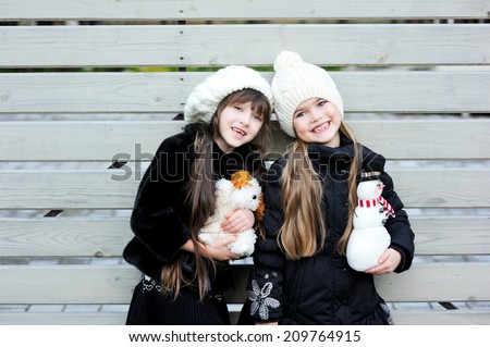 Two adorable kid girl friends in white hats and pattern black jackets has fun outdoor with Christmas toys at the winter day