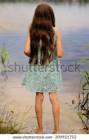 Back view of kid girl in the dress with beauty long hair stays in the water in the river at the evening
