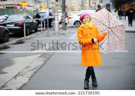 Adorable, elegant school aged kid  girl wearing orange  coat, yellow scarf and pink hat, and boots holding colorful umbrella walking in the city street autumn  day