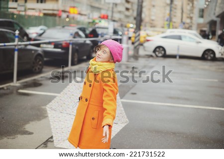 Adorable, elegant school aged kid  girl wearing orange  coat, yellow scarf and pink hat, and boots holding colorful umbrella walking in the city street autumn  day. Matte colors edit