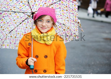 Adorable, elegant school aged kid  girl wearing orange  coat, yellow scarf and pink hat, and boots holding colorful umbrella walking in the city street autumn  day