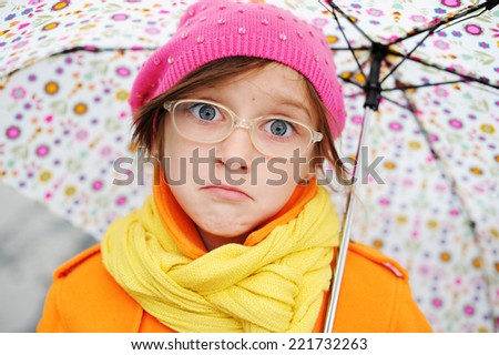 Adorable, elegan sad school aged kid  girl wearing orange  coat, yellow scarf and pink hat, and boots holding colorful umbrella walking in the city street autumn  day