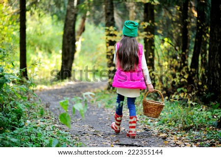 Adorable kid girl in pink vest, white sweater, green hat and boots with basket picking mushrooms in autumn forest at sunny day