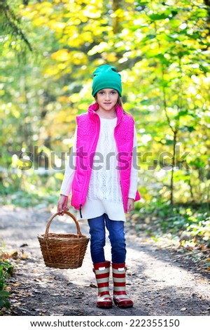 Adorable kid girl in pink vest, white sweater, green hat and boots with basket picking mushrooms in autumn forest at sunntyday