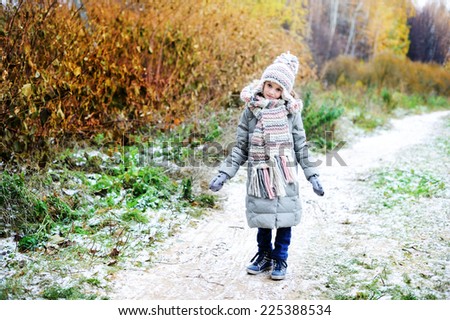 Adorable kid girl in grey coat hat and scarf walking in the fall forest with first snow