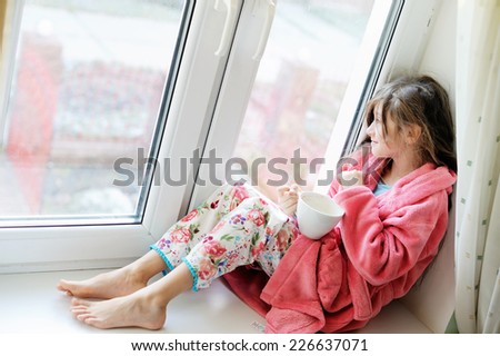 Beautiful little girl in cozy pink bathrobe drinking tea from white  cup on the window