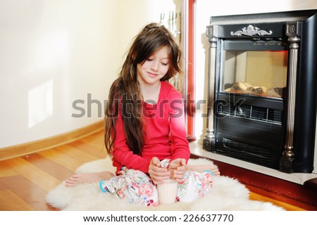 Adorable kid girl in pajama sitting near fireplace with glass of milk