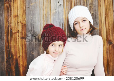 Cute kid girls of 7 and 12 years old wearing knitted trendy winter hats and sweaters posing over wood wall