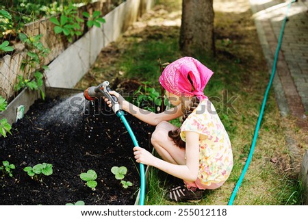 Adorable brunette kid girl, watering the plants, from hose spray in the garden at the backyard of the house on a sunny summer evening
