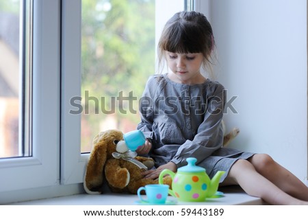 Little girl with dark hairs in gray dress with tea and bunny sitting near the window