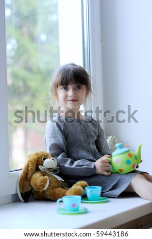 Little girl with dark hairs in gray dress with tea and bunny sitting near the window