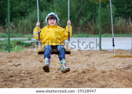 Adorable toddler girl in yellow rain coat and colorful rain boots playing on playground on swing