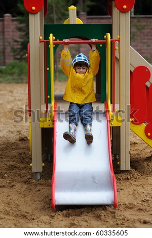 A cute young girl in yellow rain coat and colorful rain boots playing on a slide in a park