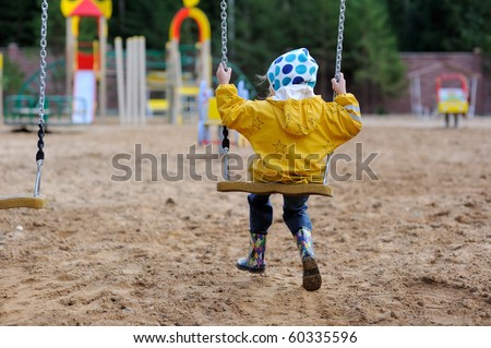 Back view of small girl in yellow rain coat and colorful rain boots playing on playground on swing