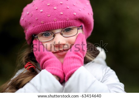 Adorable small girl in bright pink hat with long dark hair and glasses with her hand in gloves near the face