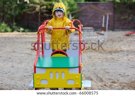 Adorable toddler girl in yellow rain coat playing on playground in playground car