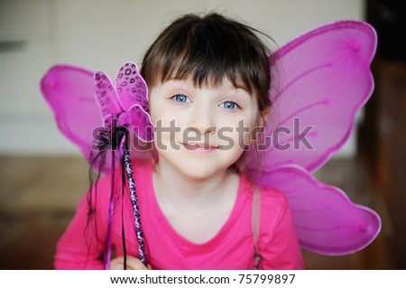 Beauty little girl with dark hair and blue eyes in pink shirt ans pink fairy wings with magic wand looks into the camera