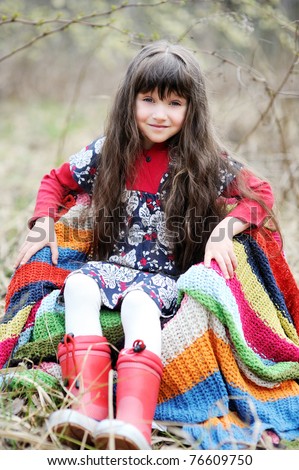 Adorable brunette little girl with very long hair in colorful dress and  red rain boots in the field with colorful stripe blanket