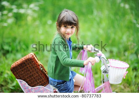 Adorable little brunette girl rides her bike with Picnic basket in the green summer field