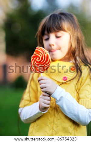 Portrait of cute little girl eating big lollipop on a stick outdoors, focus on lollipop