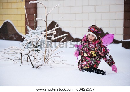 Adorable little girl in colorful snow suit with fairy wings and magic wand plays outdoors in snowfall