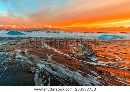 Surf on the black beach with black sand and blue floes at orange sunset  background. South of Iceland.