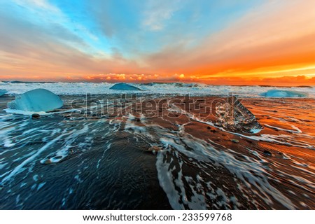 Surf on the black beach with black sand and blue floes at orange sunset  background. South of Iceland.