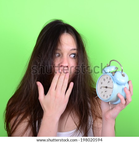Portrait of sleepy young female in chaos holding clock against green background - Running late concept