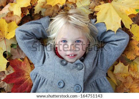 Young blond girl in gray coat laying in colorful maple leaves. Child portrait outdoors.