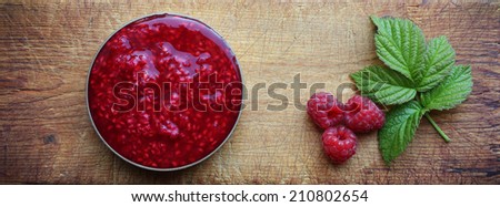 Raspberry jam, three berries and a raspberry leaf at old wooden cutting board.