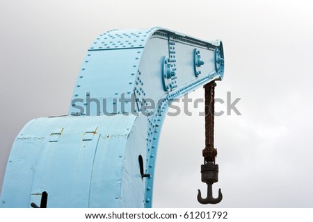 Old blue crane with rusty chain and hook on harbor with cloudy sky
