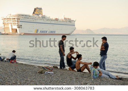 Kos, Greece - August 14, 2015: Greece-Europe-Migrants. Group of young migrants sitting of the beach and cutting the hair on Greece island Kos in front of ferry Eleftherios Venizelos.