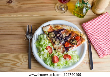 Overhead shot of a vegetarian pizza and salad, with cutlery, napkin, wine and condiments, set out on a pine wooden slatted table.