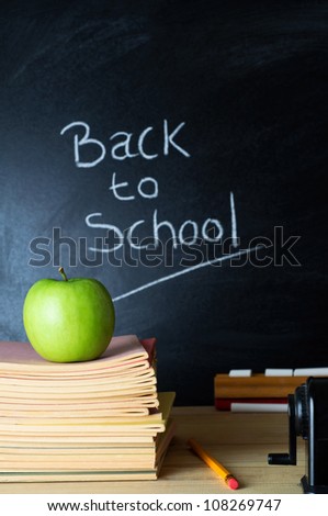 Teacher\'s desk with a pile of books, an apple and other equipment.  The words \'Back to School\' written in chalk on the blackboard in soft focus background.