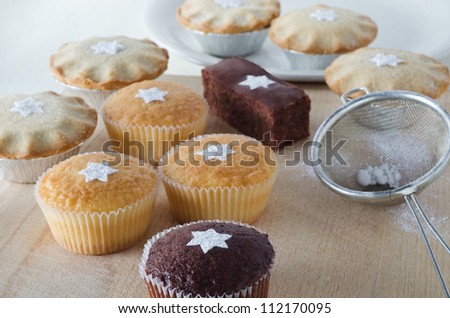 A group of Christmas cakes and mince pies, decorated with star shapes from sifted icing sugar, on a wooden chopping board with plate in background, and sifter with scattered icing sugar to the right.