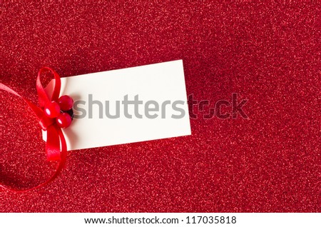 A blank Christmas gift tag, decorated with red ribbon and artificial holly berries, on a sparkly red glitter background.  Copy space on tag.