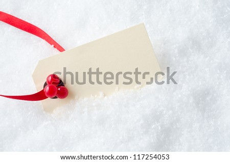 A blank Christmas gift tag, attached to a red ribbon and decorated with plastic red holly berries, nestling in artificial snow.  Copy space on cream coloured tag.